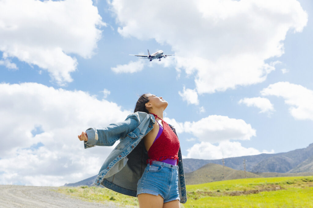 Mulher jovem de braços abertos, olhando para o céu com um avião ao fundo, em um cenário natural com montanhas e céu azul.