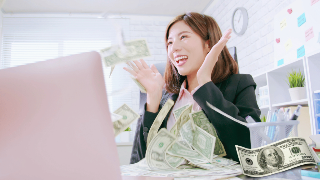 Mulher sorridente em um escritório com notas de dinheiro voando ao redor de seu computador.