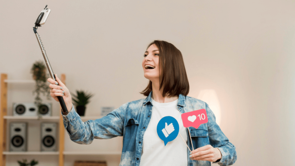 Mulher sorrindo enquanto grava um vídeo com um bastão de selfie, segurando ícones de curtidas e engajamento em um ambiente interno decorado.