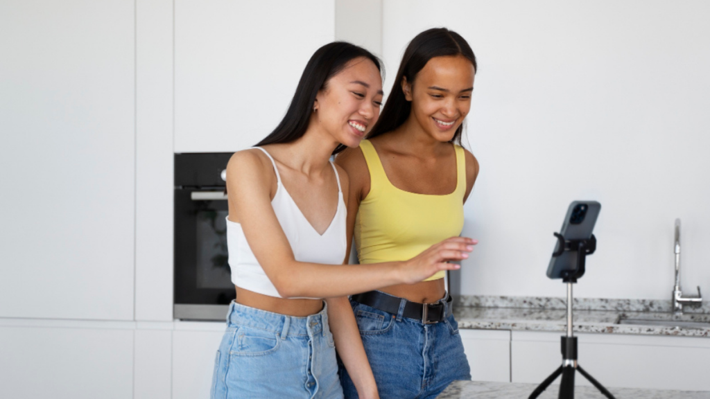 Duas jovens sorrindo enquanto gravam um vídeo em um tripé com celular, em uma cozinha moderna.