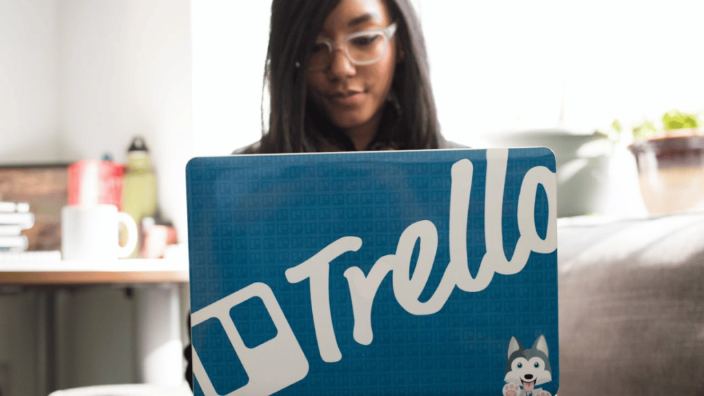 Mulher usando óculos trabalha em um laptop com um adesivo do Trello na tampa, representando organização e produtividade em um ambiente descontraído.