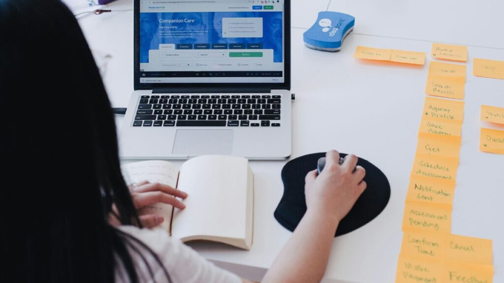 Mulher trabalhando em um laptop enquanto faz anotações em um caderno, com post-its organizados ao lado, representando um planejamento estratégico.