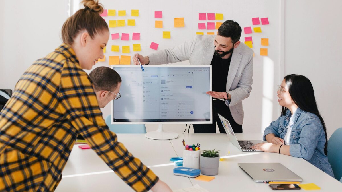 Grupo de profissionais em uma reunião de trabalho, analisando dados em uma tela de computador, com anotações em post-its ao fundo.