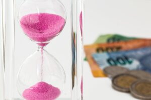 Ampulheta com areia rosa ao lado de notas e moedas, representando a relação entre tempo e dinheiro em negócios online.