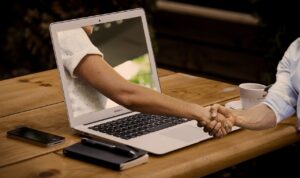 Aperto de mãos saindo de uma tela de notebook, simbolizando parcerias e negócios online firmados pela internet.
